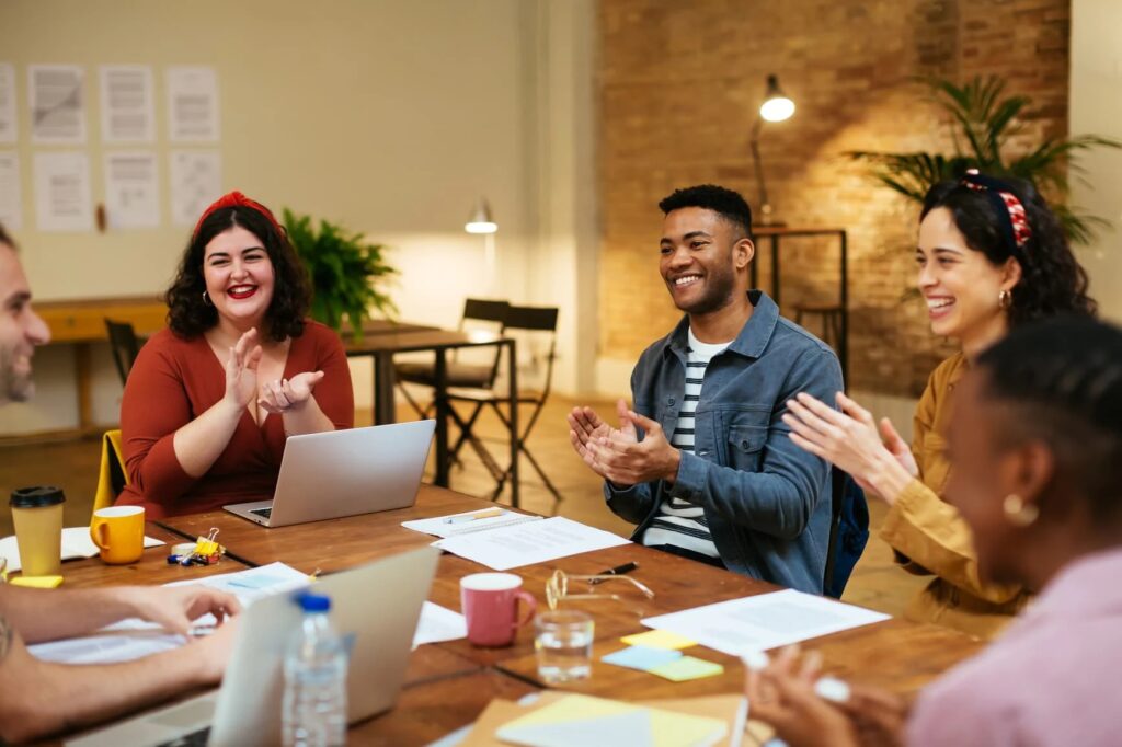 Equipe conversa e bate palmas ao redor de mesa, realizando quiz corporativo.