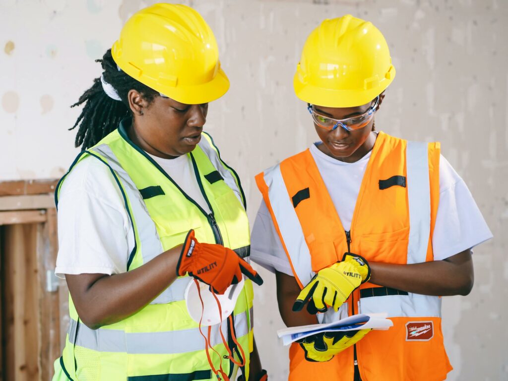 Duas mulheres em uma obra, utilizando equipamentos de proteção individual (EPIs), analisando um documento juntas, em uma cena que ilustra a prática de segurança no trabalho durante o Abril Verde.