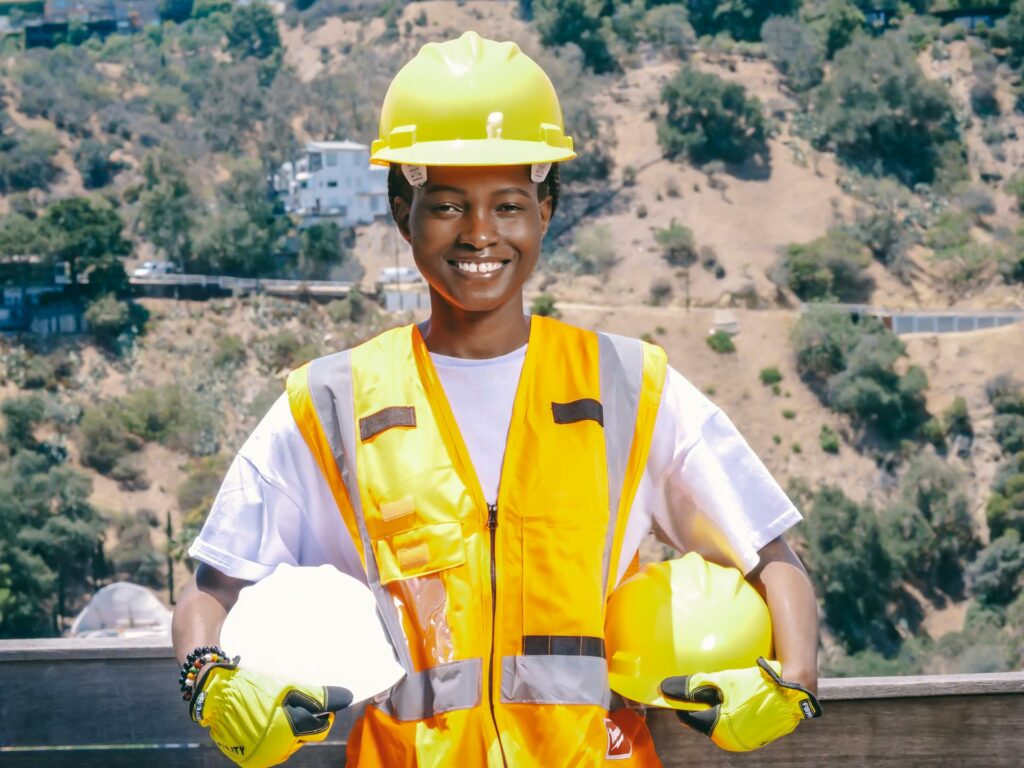 Mulher sorrindo segurando capacetes de segurança e utilizando colete de proteção, durante uma ação da campanha do Abril Verde, reforçando a importância do uso de EPIs no ambiente de trabalho.