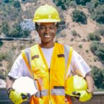 Mulher sorrindo segurando capacetes de segurança e utilizando colete de proteção, durante uma ação da campanha do Abril Verde, reforçando a importância do uso de EPIs no ambiente de trabalho.